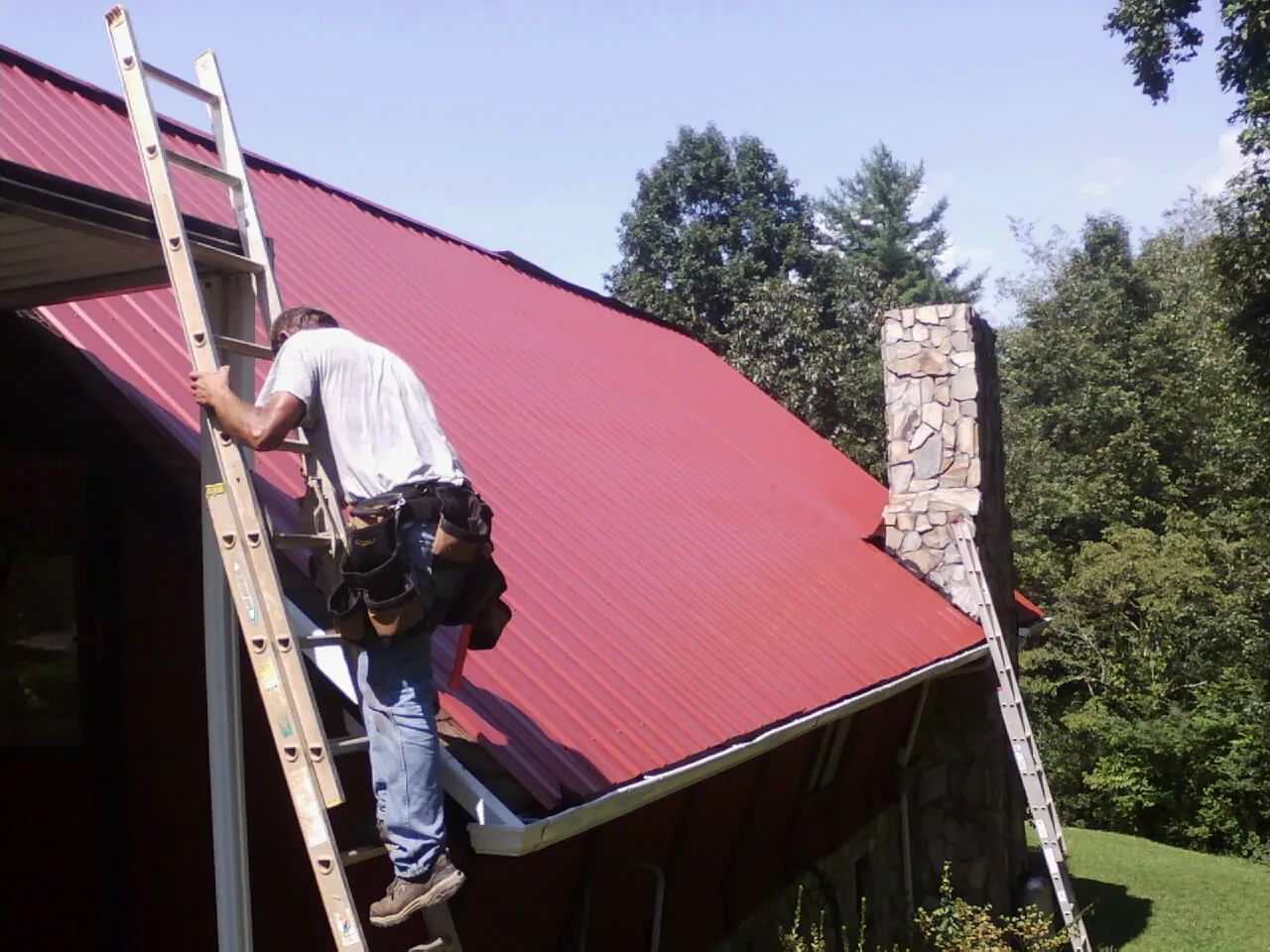 Roofer on a ladder beside a red metal roof and stone chimney — Soffit & Fascia Repair by Roofing Brothers in Cherry Hill